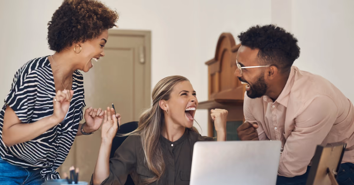 Foto de três colegas comemorando com os punhos erguidos e risadas em volta de um notebook sobre a mesa