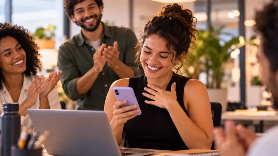 Mulher sorrindo ao olhar para o celular em uma mesa de escritório, enquanto três colegas ao redor aplaudem