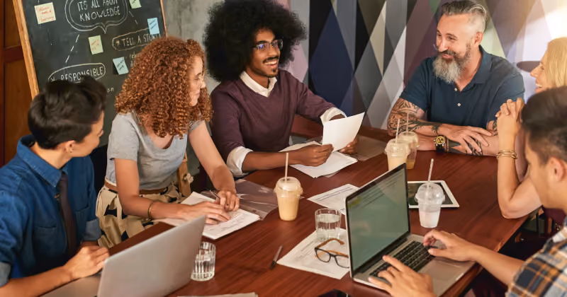Foto de seis colegas reunidos em mesa de madeira com notebooks, papéis e cafés gelados, diante de um quadro-negro com anotações
