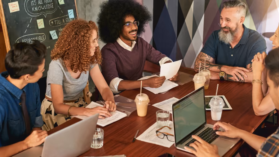 Foto de seis colegas reunidos em mesa de madeira com notebooks, papéis e cafés gelados, diante de um quadro-negro com anotações