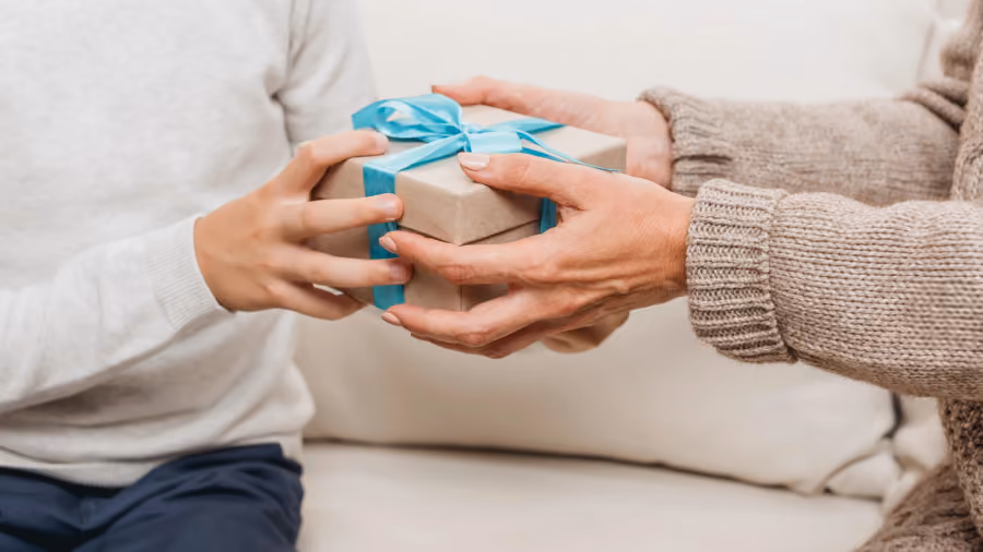 Foto em close de duas pessoas segurando juntas uma caixa de presente de papel kraft com laço azul, sentadas em um sofá