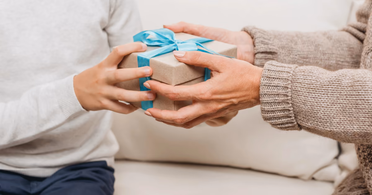 Foto em close de duas pessoas segurando juntas uma caixa de presente de papel kraft com laço azul, sentadas em um sofá