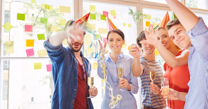 Foto de cinco pessoas com chapéus de festa e taças de espumante erguendo as mãos entre serpentinas, diante de parede com post-its