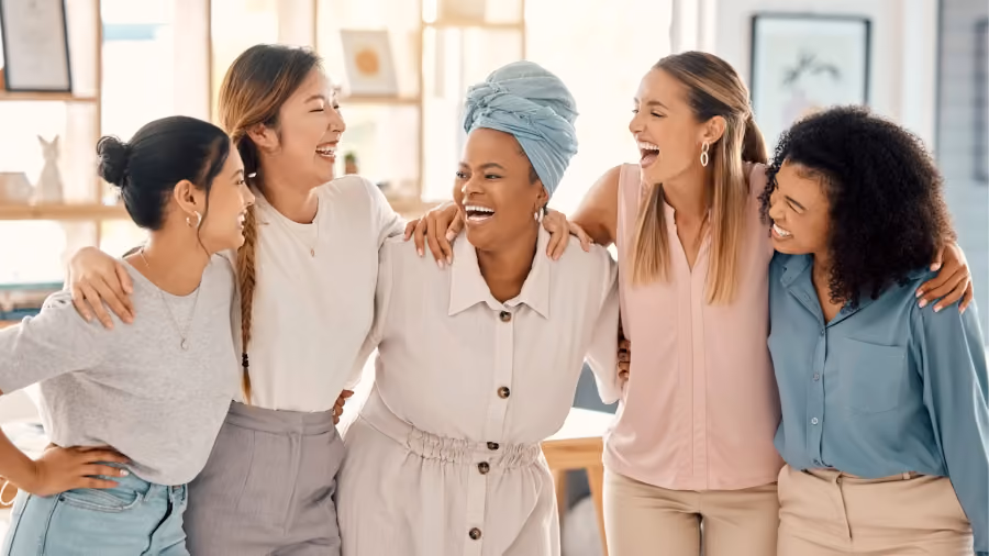 Foto de cinco mulheres abraçadas e rindo juntas em um escritório claro, ao lado de uma mesa de madeira