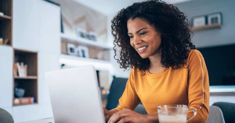 Foto de mulher de blusa amarela sorrindo enquanto digita no notebook em casa, com um copo de café ao lado