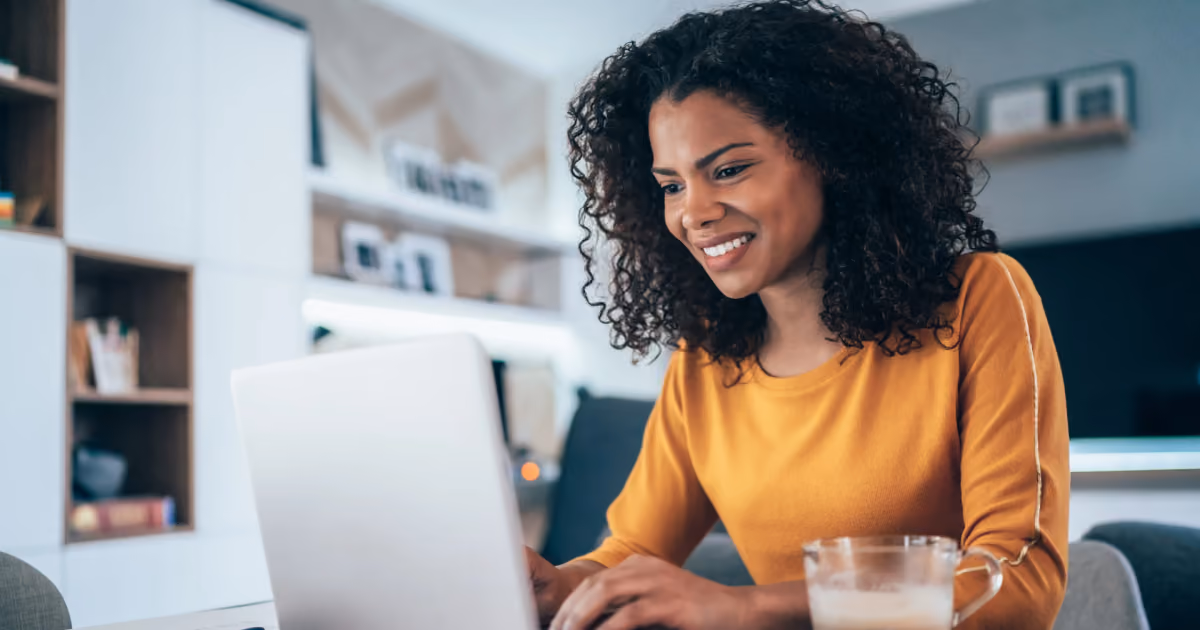 Foto de mulher de blusa amarela sorrindo enquanto digita no notebook em casa, com um copo de café ao lado