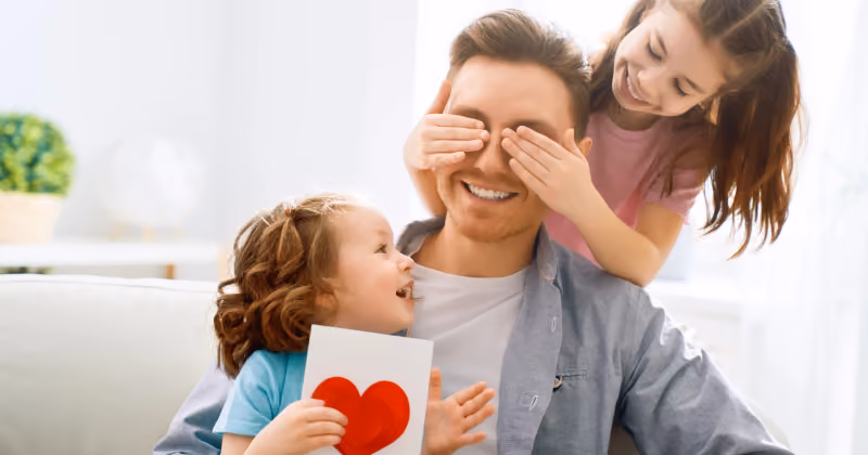 Foto de duas meninas com o pai no sofá: uma cobre os olhos dele por trás e a outra segura um cartão com coração vermelho