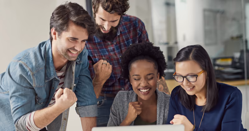 Foto de equipe de quatro pessoas olhando a tela de um notebook e vibrando com o resultado, punhos erguidos e sorrisos