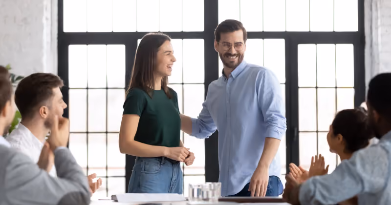 Foto de uma mulher e um homem em pé numa sala de reunião enquanto colegas sentados à mesa aplaudem