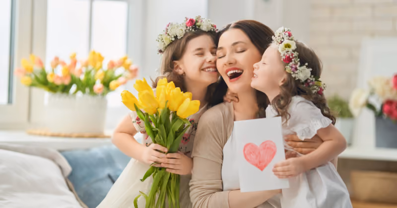 Foto de uma mãe abraçada por duas filhas com coroas de flores, segurando tulipas amarelas e um cartão com coração desenhado