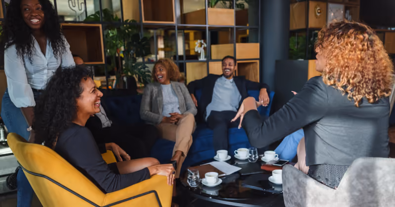 Foto de seis colegas rindo e conversando em poltronas de um lounge de escritório, com xícaras de café na mesa de centro
