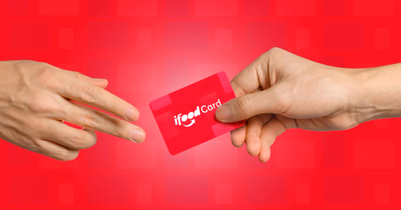 Foto de uma mão entregando a outra um cartão vermelho com a marca iFood Card, sobre fundo vermelho