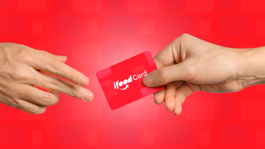 Foto de uma mão entregando a outra um cartão vermelho com a marca iFood Card, sobre fundo vermelho