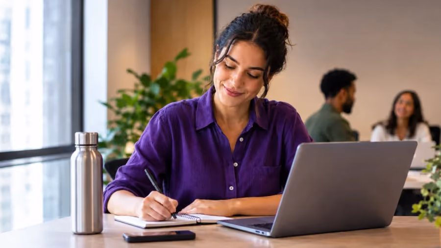 Mulher de camisa roxa escrevendo em um caderno ao lado de um notebook em uma mesa de escritório, com dois colegas ao fundo