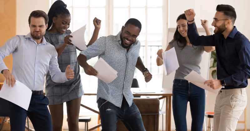 Foto de cinco pessoas comemorando de pé no escritório, com papéis nas mãos e punhos erguidos