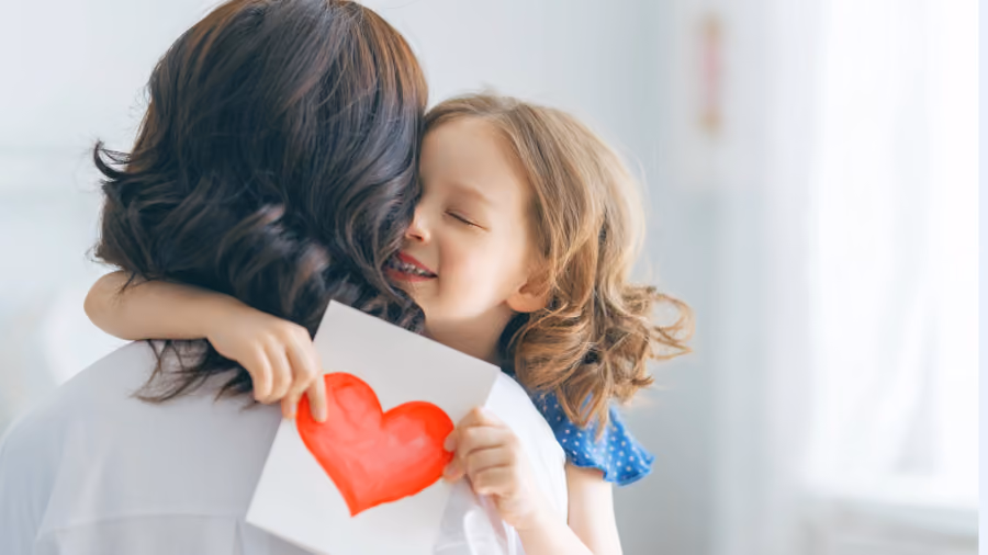 Foto de menina abraçando a mãe com os olhos fechados enquanto segura um cartão com um coração pintado em vermelho