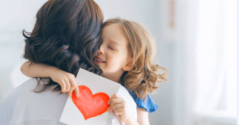 Foto de menina abraçando a mãe com os olhos fechados enquanto segura um cartão com um coração pintado em vermelho