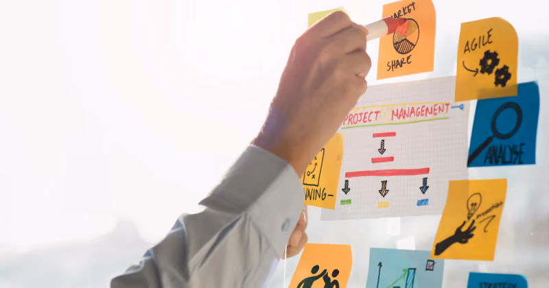 Foto de mão colando post-it em parede de vidro coberta de anotações e diagramas com palavras como project management, agile e strategy
