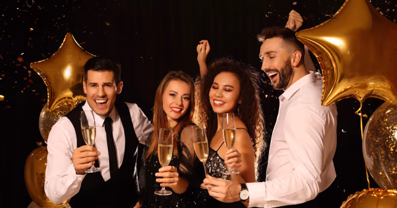 Foto de duas mulheres e dois homens brindando com taças de espumante, cercados por balões dourados em forma de estrela