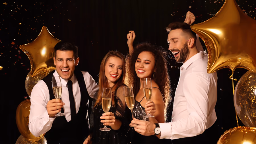 Foto de duas mulheres e dois homens brindando com taças de espumante, cercados por balões dourados em forma de estrela