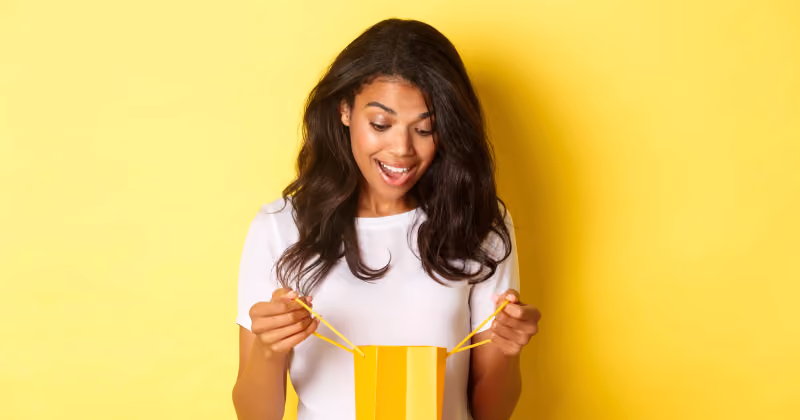 Foto de jovem de camiseta branca sobre fundo amarelo, sorrindo ao abrir e olhar dentro de uma sacola de presente
