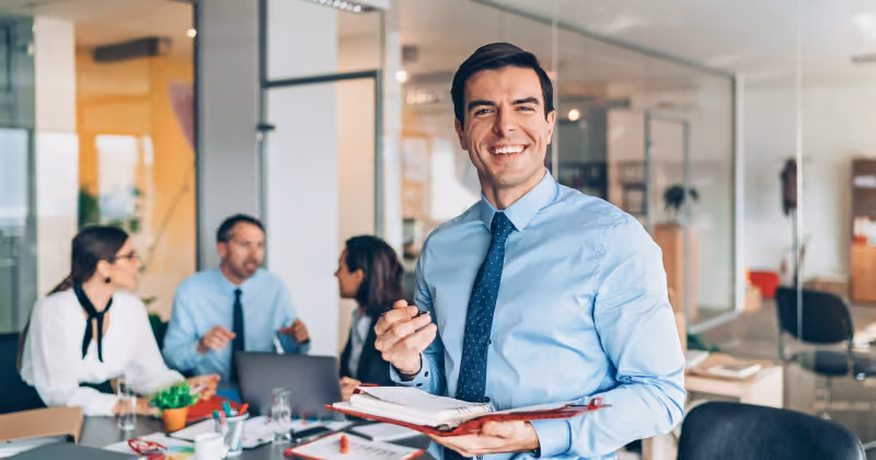Foto de homem de camisa azul e gravata sorrindo com uma pasta vermelha aberta nas mãos, com colegas reunidos ao fundo