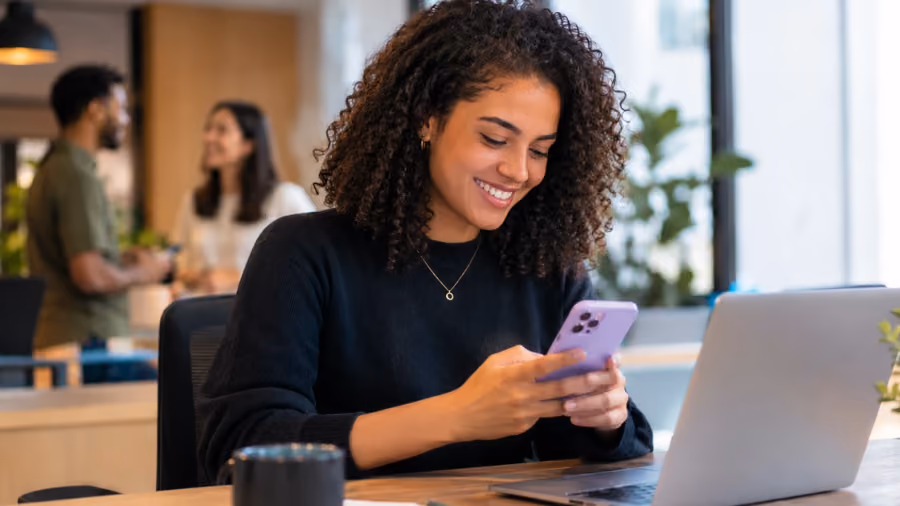 Mulher sorrindo ao olhar para o celular em uma mesa de escritório, com um notebook ao lado e dois colegas conversando ao fundo