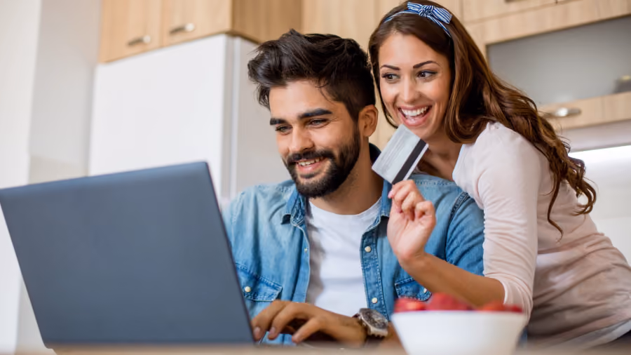 Foto de casal sorrindo diante de um laptop na cozinha, com a mulher segurando um cartão na mão