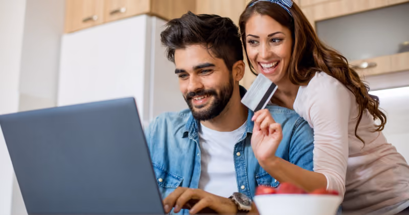 Foto de casal sorrindo diante de um laptop na cozinha, com a mulher segurando um cartão na mão