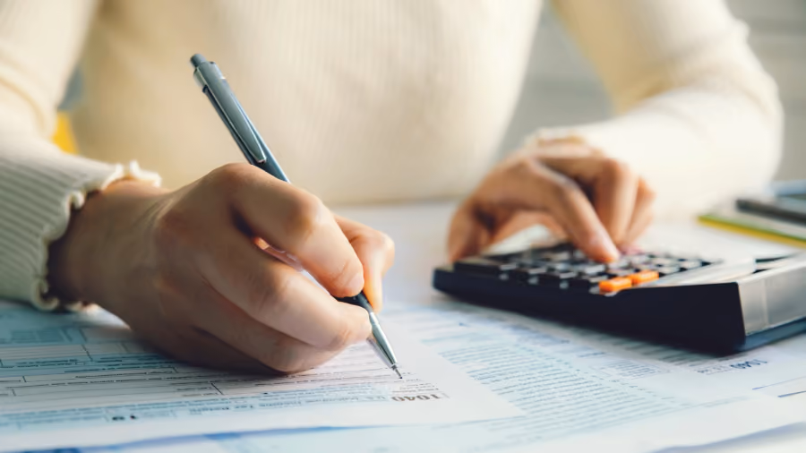 Foto em close de mãos preenchendo um formulário fiscal com caneta enquanto operam uma calculadora sobre a mesa