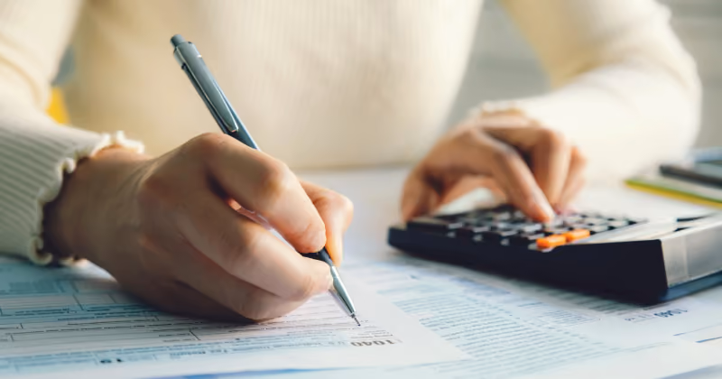 Foto em close de mãos preenchendo um formulário fiscal com caneta enquanto operam uma calculadora sobre a mesa