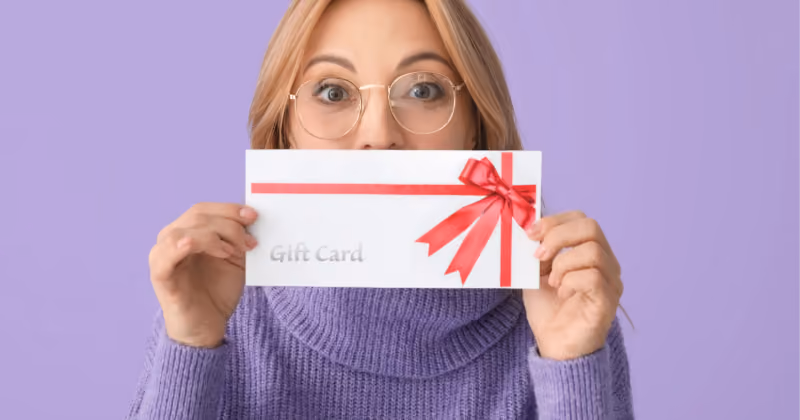 Foto de mulher de óculos segurando diante do rosto um envelope branco com laço vermelho e a inscrição Gift Card