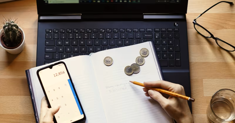 Foto vista de cima de mesa com notebook, caderno aberto com moedas, celular na calculadora e mão segurando lápis