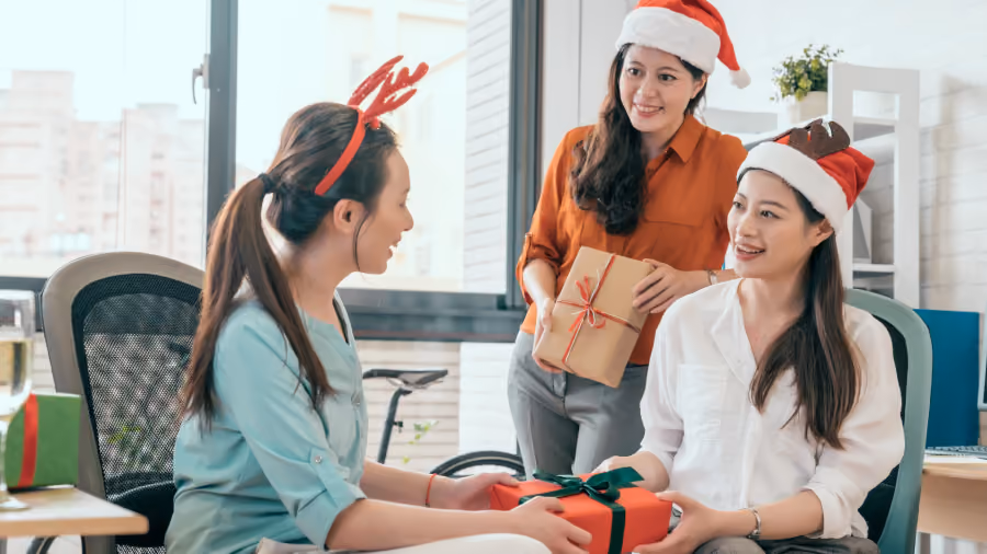 Foto de três colegas de escritório com gorros de Papai Noel e tiara de rena trocando presentes embrulhados